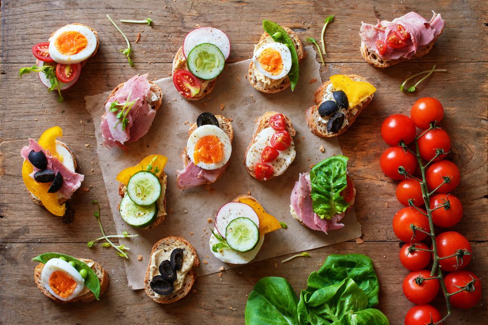 Variedad de canape con hummus, jamón y queso, huevos y verduras frescas en mesa de madera.